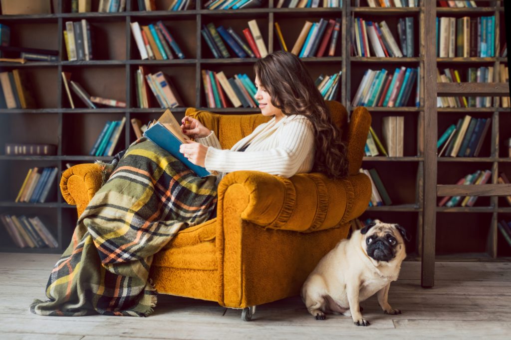 Woman reading in an armchair with a plaid blanket covering her legs. A pug dog is sitting next to the chair.